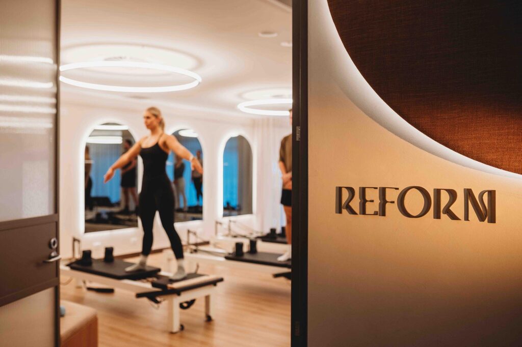 Woman doing pilates in a bright modern interior, warm earthy tones, timber floor, arched mirrors, funky sign, fabric panel